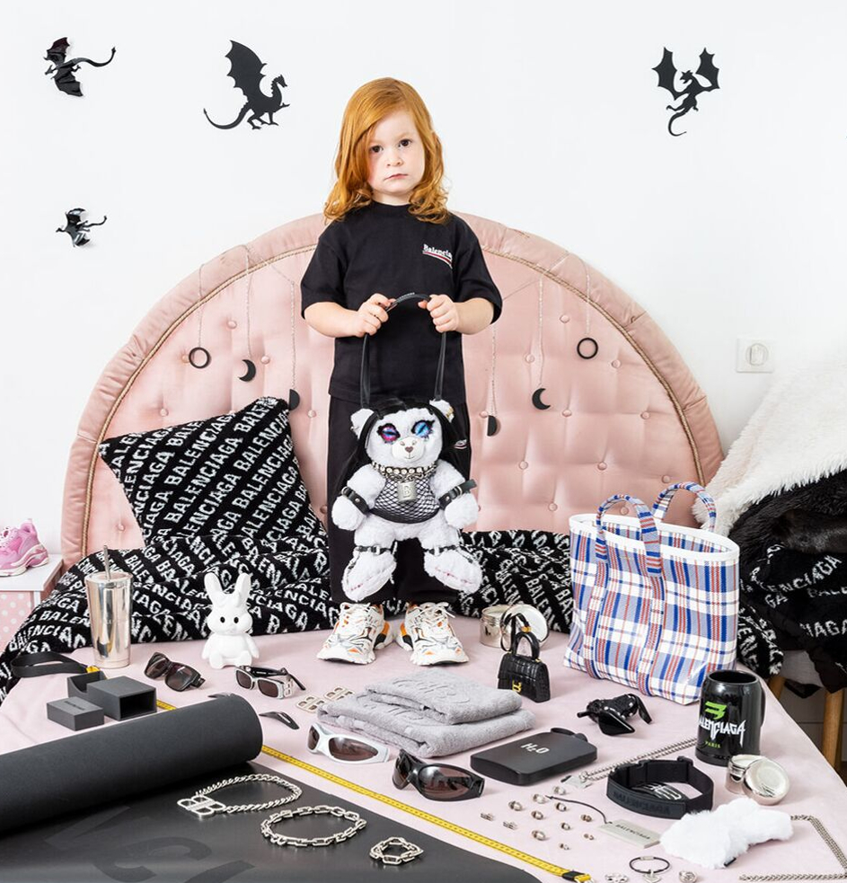 A young redhead child is pictured holding a teddy bear with bondage-themed apparel 